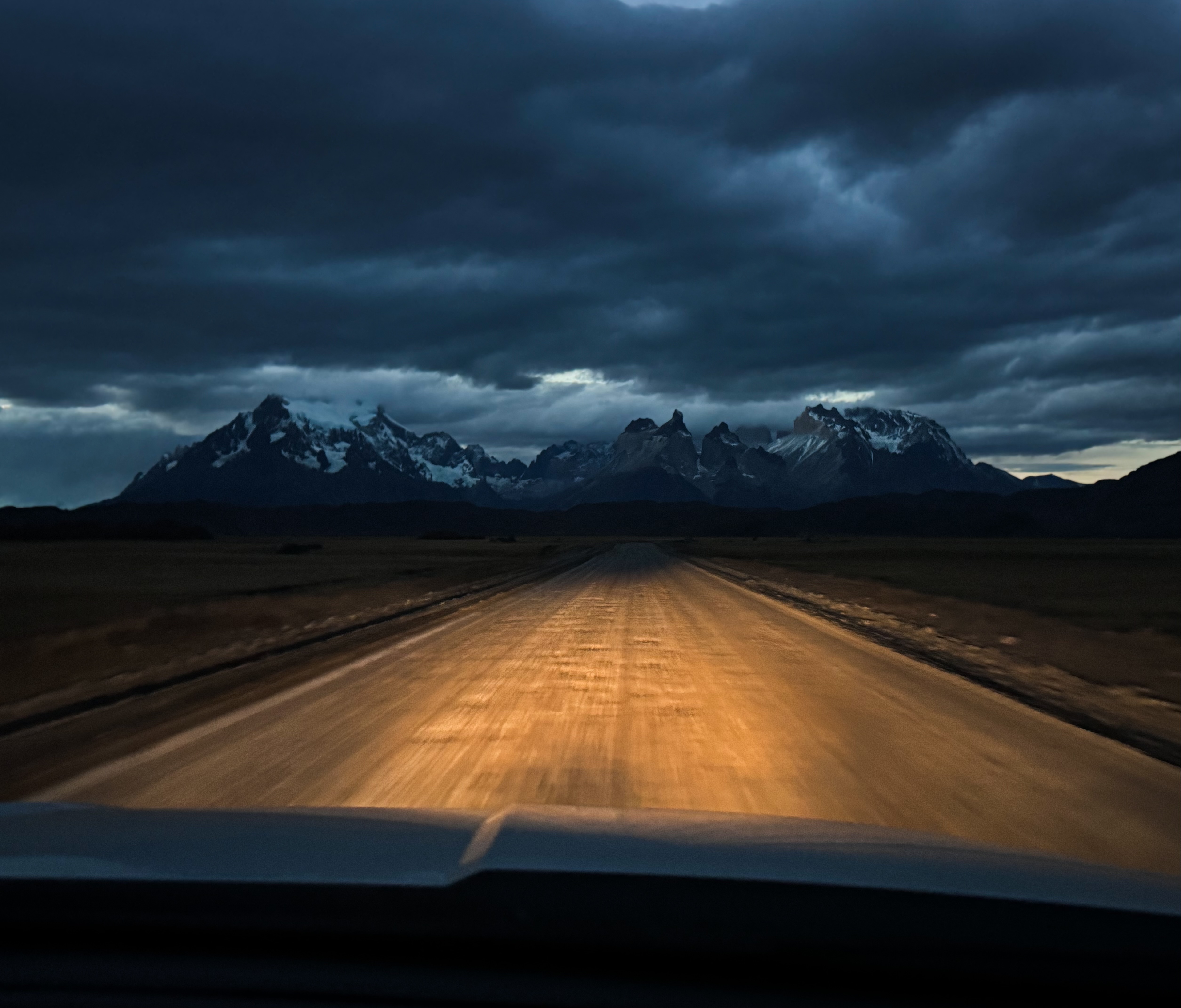 A car lights up a bumpy road paving the way to the peaks of Patagonia, showing the traces of the famous Patagonia logo, dimly lit by the setting sun